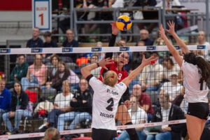 Marian Mischo von den BayerVolleys Leverkusen, im roten Trikot mit der Nummer 9, holt über dem Netz zum Angriffsschlag aus. Der Ball schwebt über ihrer Hand. Zwei Spielerinnen des VfL Oythe in Weiß springen mit gestreckten Armen zum Block, links die Numme