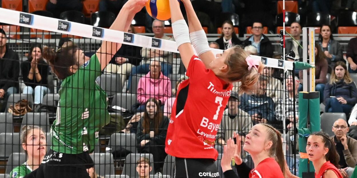 BayerVolleys-Spielerin Alice Turmovich springt im roten Trikot am Netz zum Block gegen die Zuspielerin des TV Planegg-Krailling; im Hintergrund verfolgen Zuschauerinnen und Zuschauer die Szene in der Ostermann-Arena.