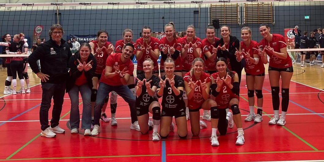 Die Spielerinnen und das Trainerteam der BayerVolleys posieren nach dem WVV-Pokalsieg in roten Trikots auf dem Volleyballfeld.