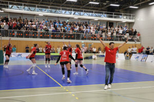 Spielerinnen der BayerVolleys Leverkusen jubeln nach dem 3:1-Auswärtssieg im Rheinland-Derby bei DSHS SnowTrex Köln; im Vordergrund feiert ein Mitglied des Leverkusener Teams mit erhobenen Armen, im Hintergrund stehen die Zuschauer auf den Rängen.