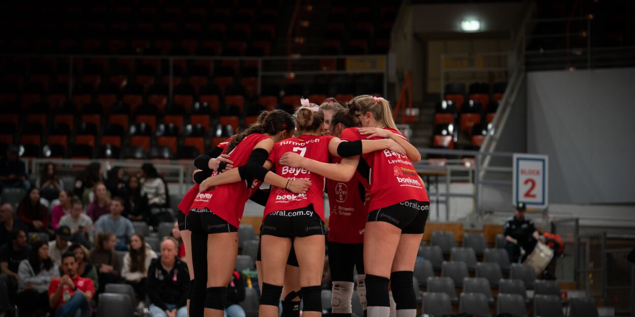 Die Spielerinnen der BayerVolleys Leverkusen stehen in roten Trikots eng im Kreis und halten sich in den Armen. Im Hintergrund sitzen einige Zuschauer auf der Tribüne, der Rest der Halle ist leer und dunkel.