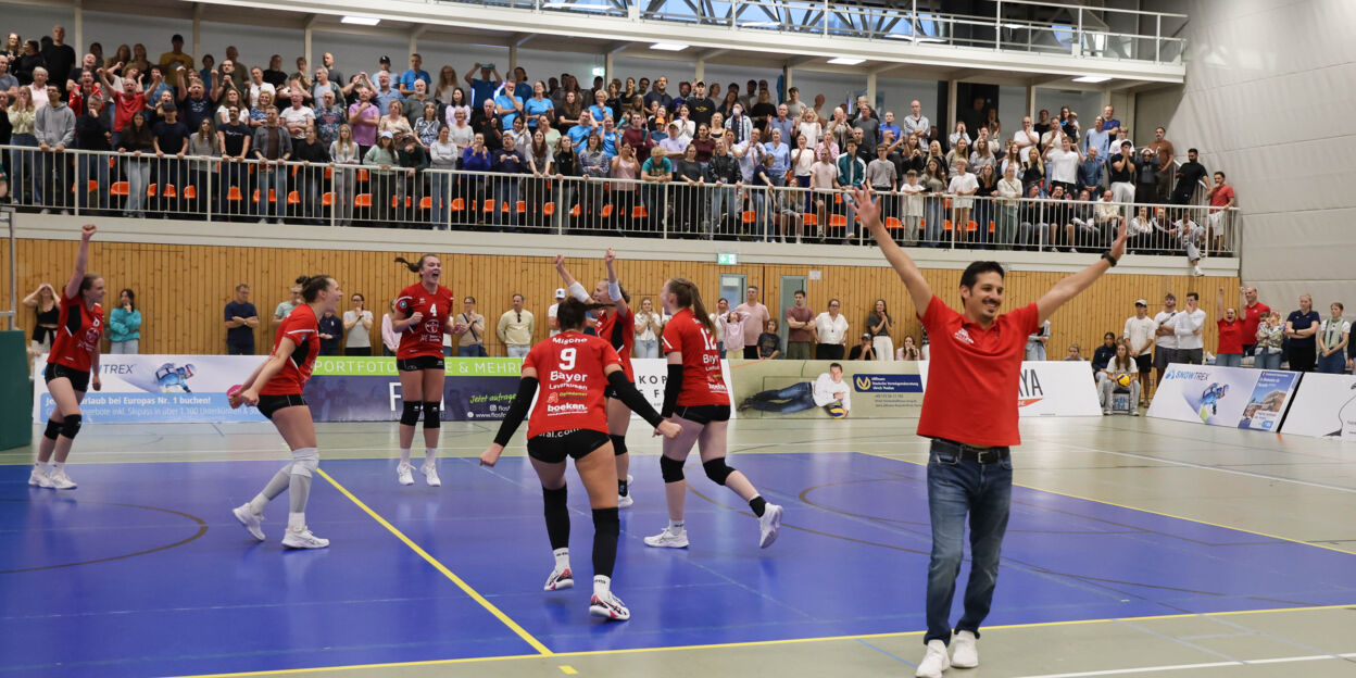 Spielerinnen der BayerVolleys Leverkusen jubeln nach dem 3:1-Auswärtssieg im Rheinland-Derby bei DSHS SnowTrex Köln; im Vordergrund feiert ein Mitglied des Leverkusener Teams mit erhobenen Armen, im Hintergrund stehen die Zuschauer auf den Rängen.